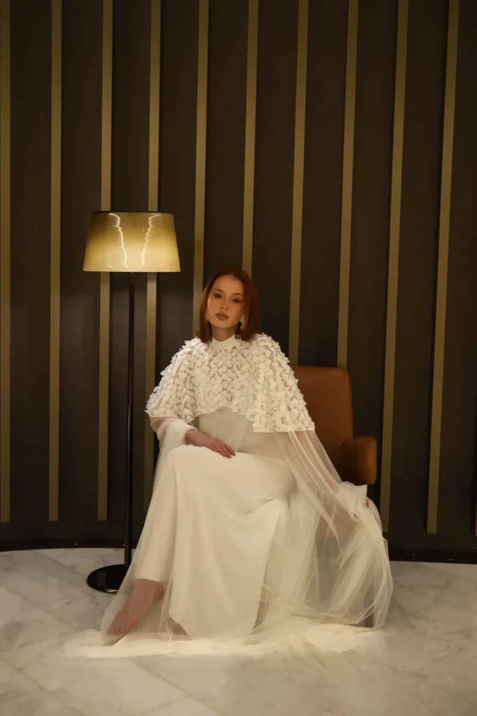 A woman in a long white dress sits on a brown chair next to a floor lamp against a dark wall with vertical gold stripes.